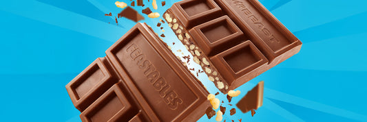 A milk chocolate bar labeled “FEASTABLES” and “MRBEAST” is broken in half, revealing a crunchy, textured filling, with chocolate pieces and crumbs scattered on a blue background.