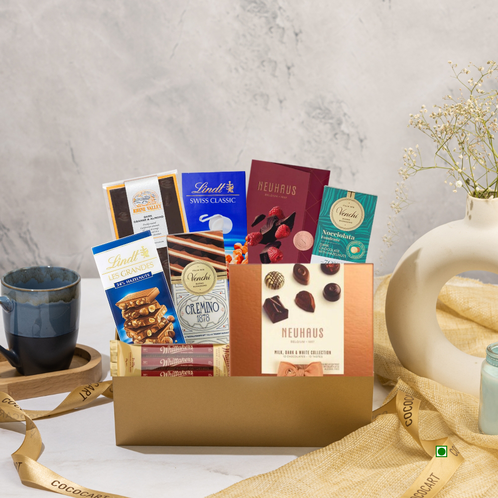 The Sweet Selection Giftbox by Gift Hampers, featuring an exquisite selection of international chocolate bars and packages, is elegantly presented on a table, adorned with a ribbon, teacup, and a vase filled with flowers.