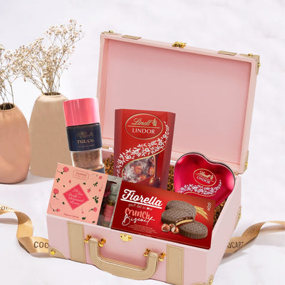 Pink Cupid's Choice Hamper gift box open displaying various Lindt Lindor chocolates, a Hazelnut Cream perfume bottle, and a potpourri bag, set against a neutral background with dry flowers.