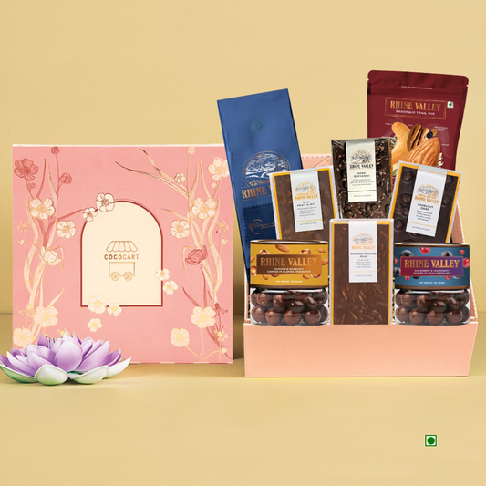 The "Feminine Finesse: Rhine Valley Edition" by Gift Hampers showcases an array of chocolates and snacks, beautifully encased in pink packaging with floral motifs. Positioned on a beige backdrop with a purple lotus, this gift promises an enchanting journey reminiscent of the Rhine Valley.