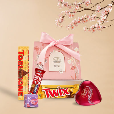 A gift assortment with chocolates including Kit Kat Chunky and Twix, and a pink gift bag with a ribbon, against a backdrop of cherry blossoms from Gift Hampers' Hugs & Kisses - Mother's Day Collection.