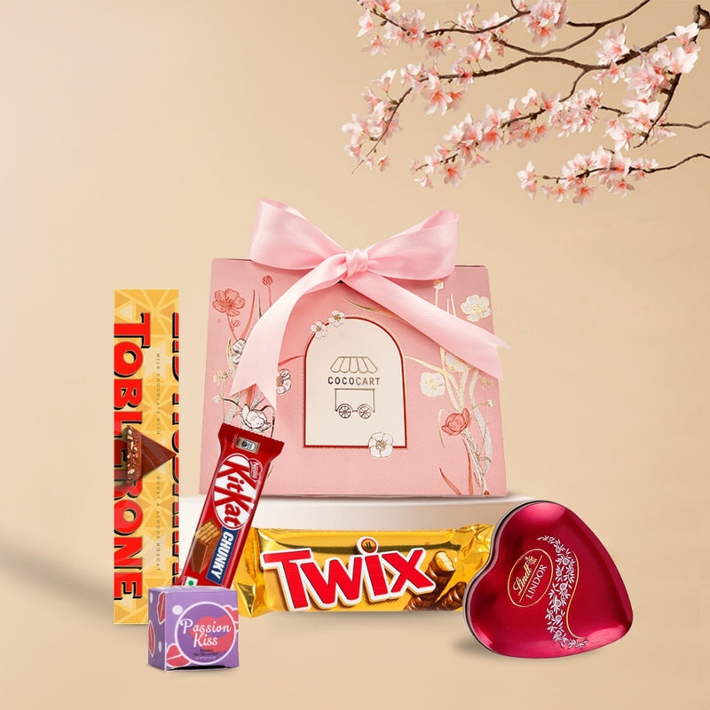 A gift assortment with chocolates including Kit Kat Chunky and Twix, and a pink gift bag with a ribbon, against a backdrop of cherry blossoms from Gift Hampers&