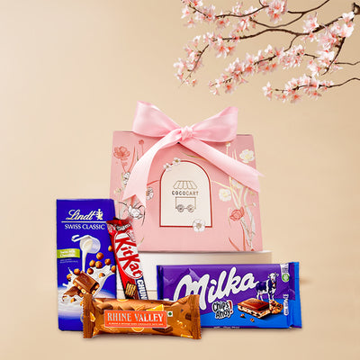 A Sweet Solace - Mother's Day Collection gift basket with assorted chocolates including Lindt Swiss Classic, Milka Chips Ahoy, and Toblerone, with a pink ribbon and floral decor by Gift Hampers.