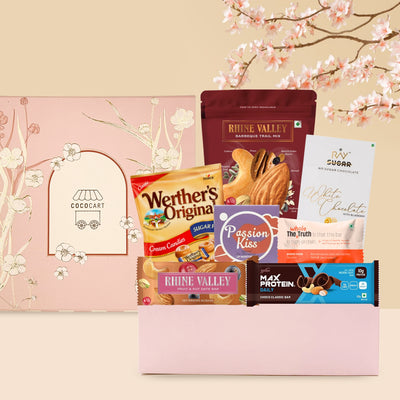 A selection of Guilt Free Indulgence - Mother's Day Collection snacks including sugar free chocolate, cookies, and protein bars displayed against a floral background with a pink notebook from Gift Hampers.
