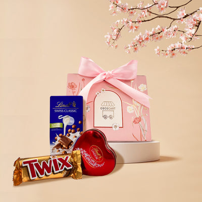 Assorted Swiss chocolates and a pink gift bag with floral design set against a beige background with cherry blossoms - Milk Chocolate Delight - Mother's Day Collection by Gift Hampers.