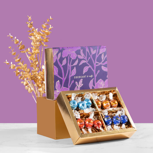 An open decorative mini gift box from The Dusk Collection by Gift Hampers showcases Lindor assorted chocolates next to a closed floral design box on a purple background. Elegantly arranged golden leaves evoke the rich Swiss heritage and tease with the promise of exquisite dark delights inside.