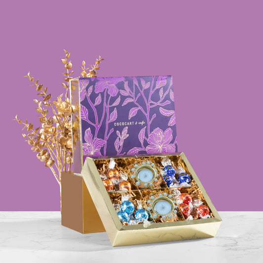 The Dusk Collection: Lindor Giftbox by Gift Hampers, featuring assorted colorful chocolates, including Lindor Milk, alongside two candles, elegantly arranged on a marble surface. The gift box lid is adorned with floral patterns and a logo. Golden decorative leaves enhance the background.