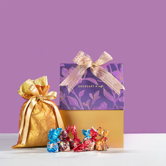 The Dusk Collection: Assorted Pyramid Giftbox from Gift Hampers features a sophisticated arrangement of a purple gift box accented with a gold bow, accompanied by an exquisite golden Potli Round pouch, and an assortment of wrapped chocolates, all elegantly displayed on a white surface against a purple backdrop.