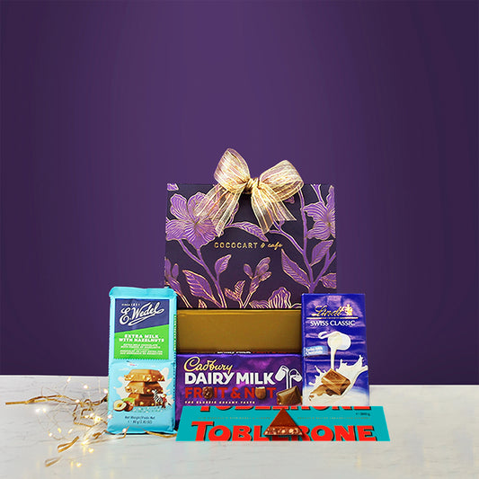 Gift Hampers presents The Dusk Collection: The Opulence Edit Pyramid Giftbox, wrapped with a gold bow and containing chocolate bars like Cadbury Dairy Milk Fruit & Nut, L.Widde Extra Milk Toffee, Lindt Swiss Classic Milk Bar, and Toblerone, all set against a luxurious purple background.