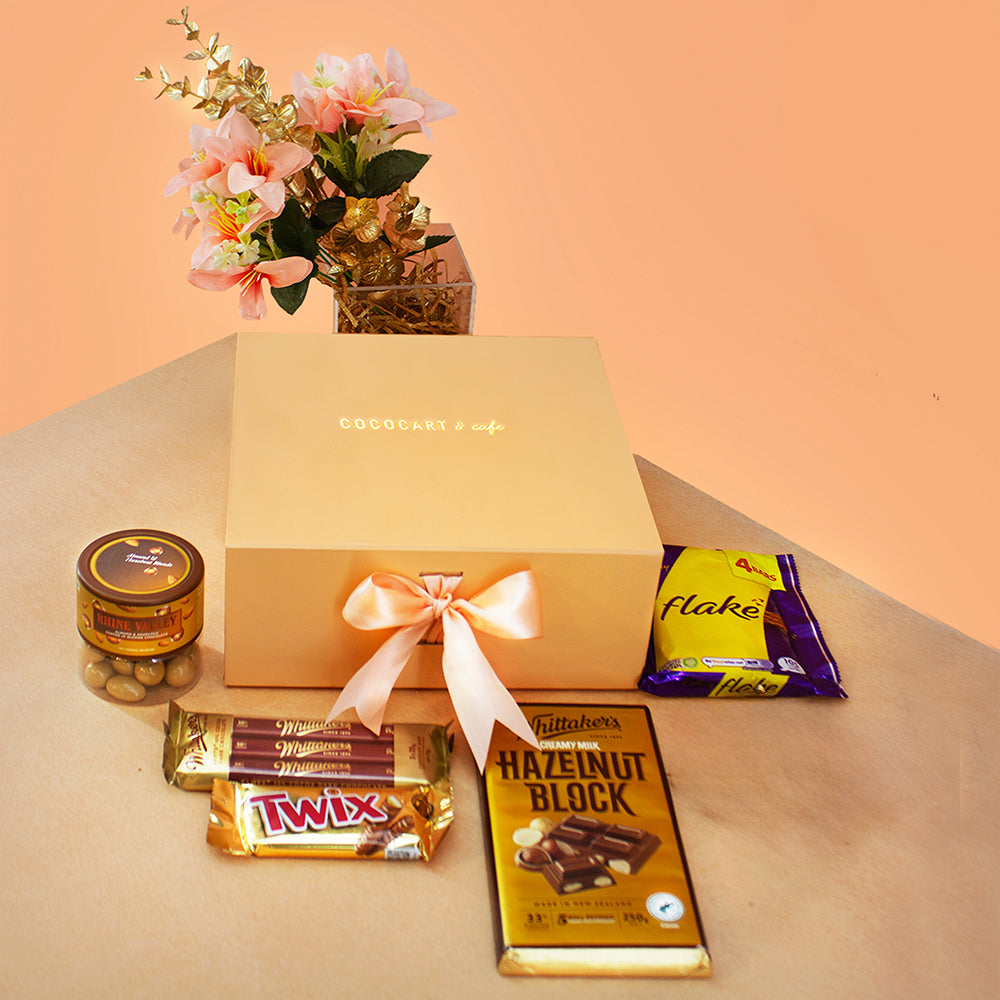 A gift box labeled "The Dawn Collection: Decadence Giftbox" from Gift Hampers, adorned with a bow, is surrounded by Cadbury Flake, Twix, and Lindt Lindor chocolates on a table, accompanied by a jar of nuts and a floral arrangement.