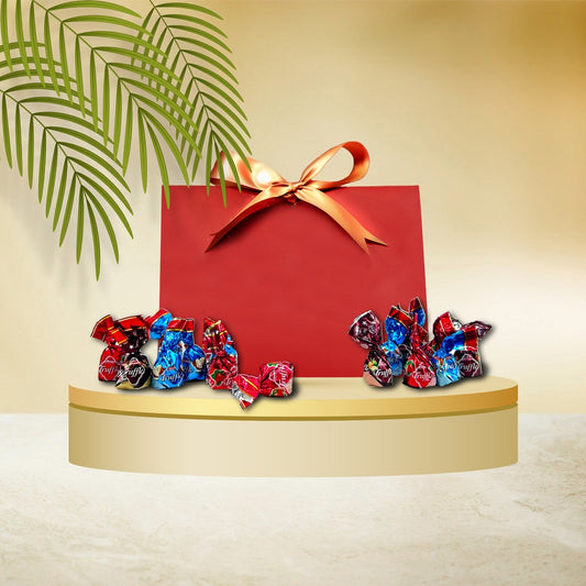 A red gift bag with a gold ribbon sits on a round platform, surrounded by assorted wrapped candies. A green plant branch extends from the top left, capturing the elegance of The Secret Santa Hamper by Gift Hampers.