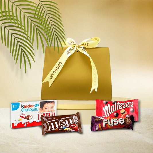 The Junior's Celebration Box by Gift Hampers, featuring Kinder Chocolate, M&M's Choco, Maltesers, and a Cadbury Fuse Chocolate Bar, sits next to a gold gift box with a ribbon against a backdrop adorned with leafy plants.