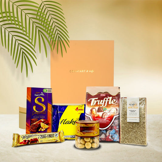 The Season’s Greetings Giftbox by Gift Hampers features Cadbury chocolates, an Almond & Hazelnut Blonde Dragees mix, truffles, cookies, a plum cake, and dried herbs on a light backdrop with a decorative plant.