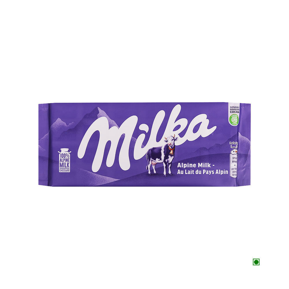 A purple Milka Milka Alpine Milk Bar 100g wrapper featuring an Alpine landscape, a cow, and text stating "Alpine Milk Chocolate - Au Lait du Pays Alpin," promises a tender taste experience.