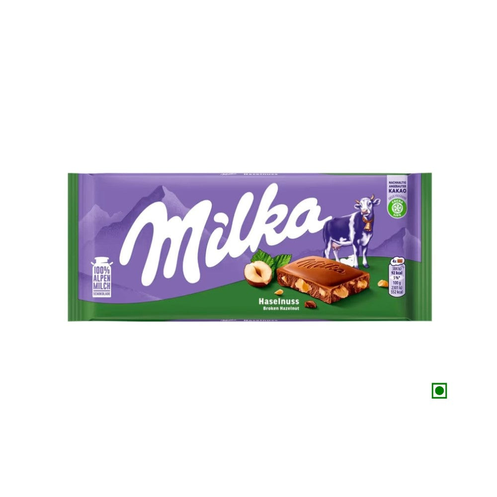 Milka Broken Hazelnut Milk Bar 100g with purple packaging featuring a cow and images of hazelnuts and chocolate squares.