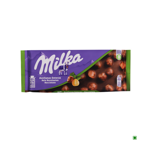 Milka Whole Hazelnut Milk Bar 100g in a purple and green wrapper, featuring the Milka logo and a cow image. This delightful German chocolate contains text in Spanish, German, and French.