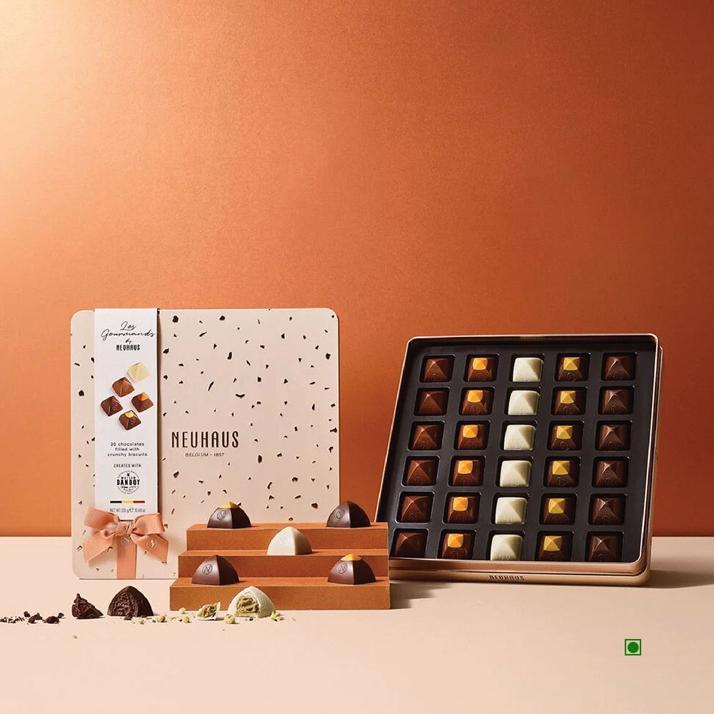 An elegant tin box houses the Neuhaus Les Gourmands 30-piece collection, presenting an array of Belgian pralines in diverse shapes and colors against a luxurious brown backdrop.