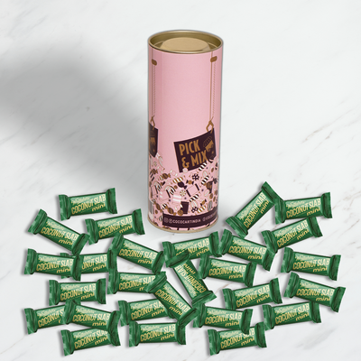A cylindrical tin of Whittakers Loose Coconut Squares Mini Slab 100/200gm chocolates surrounded by multiple green Whittakers Toasted Coconut Slab chocolate bars on a marble surface.
