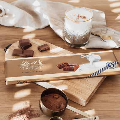 Lindt's Lindt Gold Tab Milk Chocolate 300g on a cutting board next to a cup of coffee.